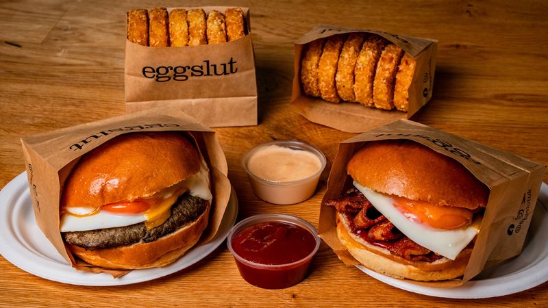 Be transported to sunny LA with eggslut's breakfast buns piled high with tasty fillings. Credit: eggslut. Image courtesy of eggslut. Two brioche buns from eggslut with a bright orange fried egg on top of bacon and a beef patty, with sauces and sides on the wooden table.