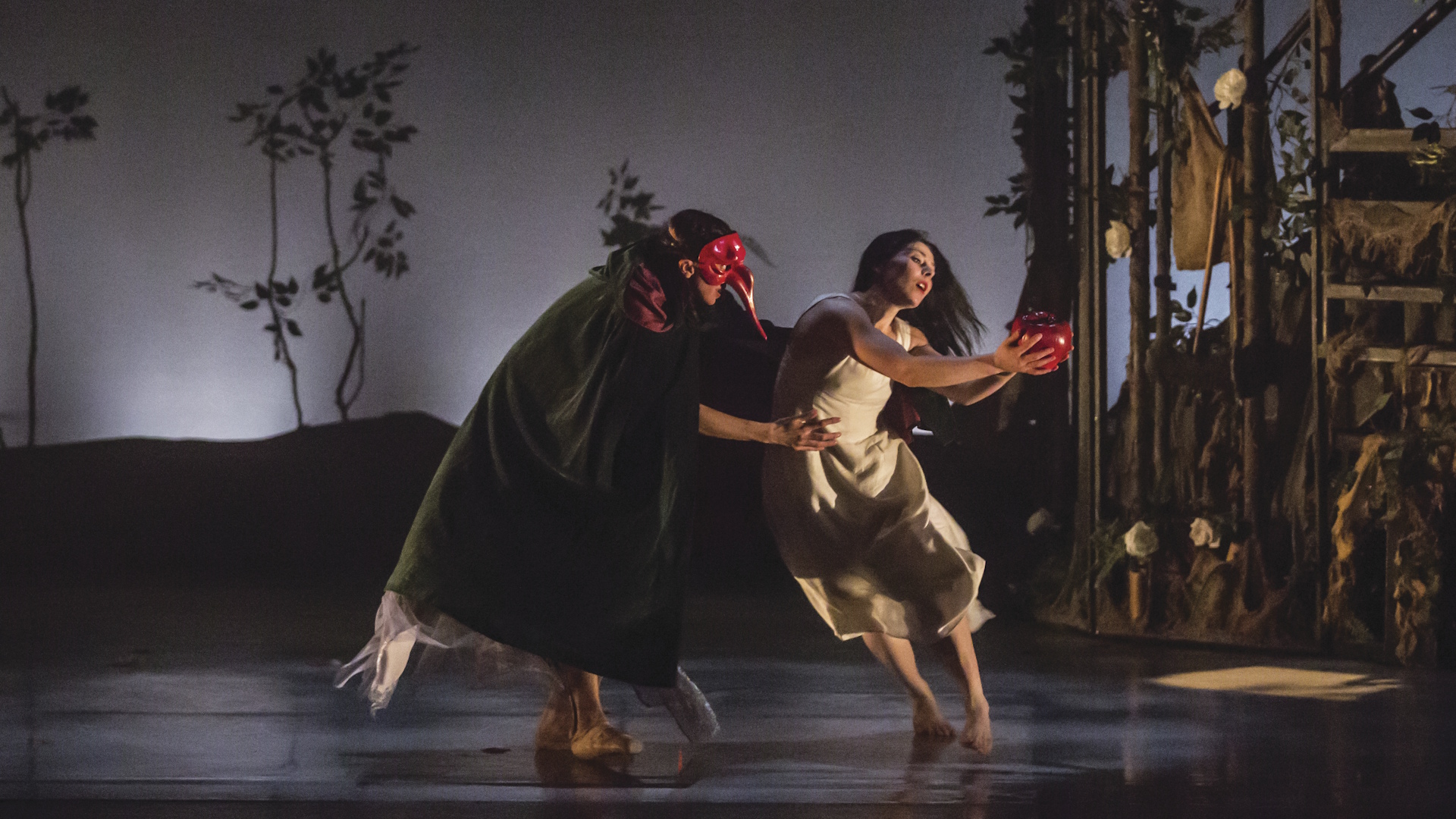 Two female dancers, one dressed in black and wearing a red mask and another one in a white dress, performing Snow White on stage.
