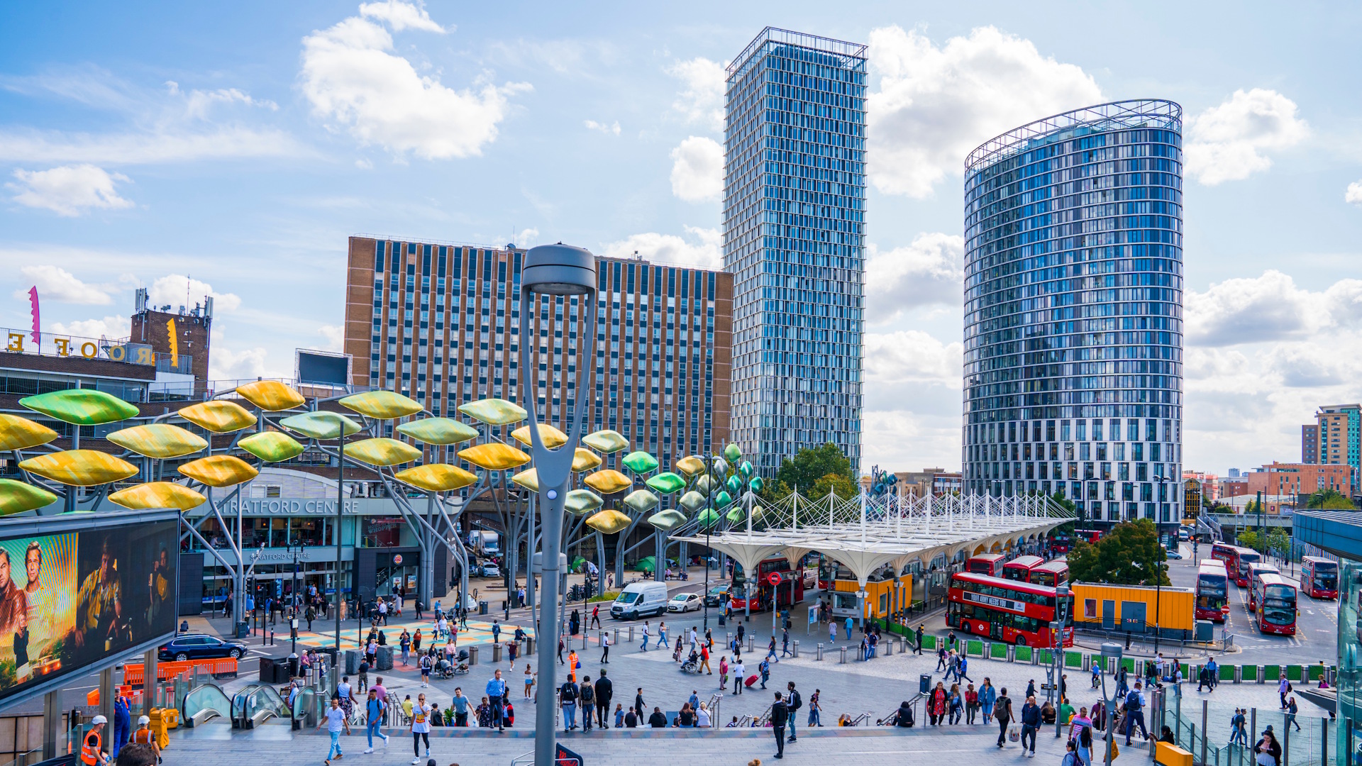 Crowds outside Stratford Station and Westfield Stratford City shopping centre, with high-rise buildings, red double-decker buses and blue skies in the background.