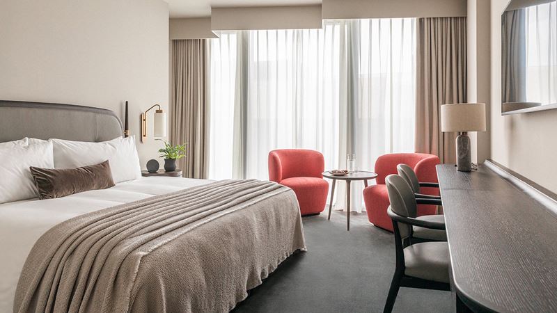 Enjoy a luxury stay featuring sleek Scandinavian interiors and plenty of style. Credit: The Stratford, Autograph Collection. Image courtesy of The Stratford, Autograph Collection. Plush hotel bedroom with double bed, white sheets, grey bed throw, curtains and carpet, beige walls and two orange armchairs.
