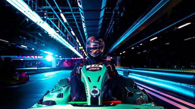 Person in the driving seat of a green Formula 1 car on an indoor racing track with dark blue lighting. 