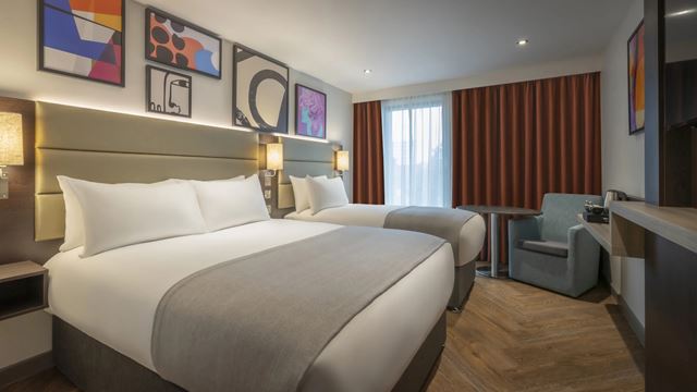 Hotel bedroom with two double beds, wooden floors, colourful artwork on the walls, orange curtains and grey and white sheets.