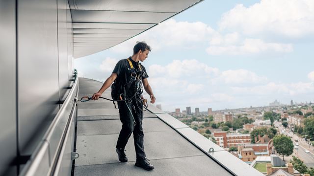 A man, attached to a railing, peers over the edge of Tottenham Hotspur Stadium, with London's skyline in the background.