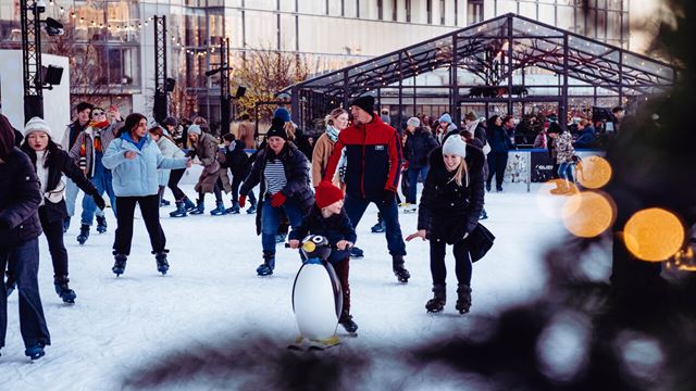Group of people skating at Glide ice rink around sparkling Christmas tree with Glass House in the background.
