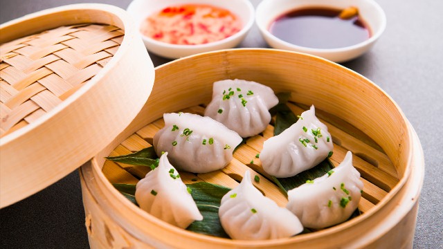 Des raviolis types dim sum aux crevettes dans un panier en bois avec des sauces à l'arrière-plan.