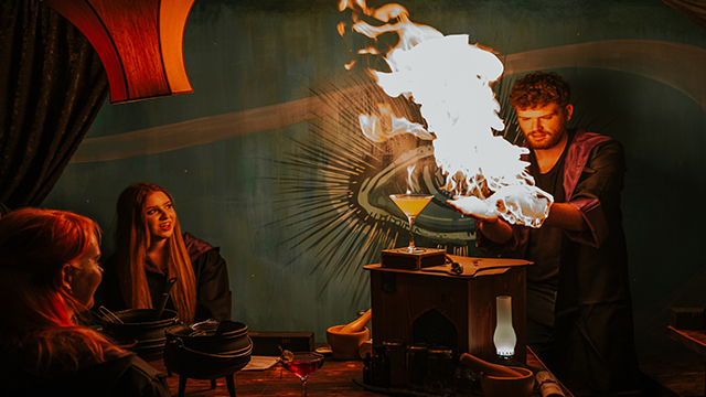 Immersiver Cocktail-Kurs zur Herstellung von Zaubertränken im Cauldron, Bild mit Genehmigung von Buyagift Der Zaubertrankmeister demonstriert die Herstellung von Cocktails mit Feuer, das seine Hände umgibt, vor zwei Frauen, die von diesem Trick sehr beeindruckt sind.