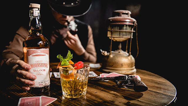Moonshine Saloon Cocktail Erlebnis, Bild mit Genehmigung von Buyagift Ein Cowboy hält eine Schnapsflasche aus dem Moonshone-Saloon in der Hand, auf dem Tisch stehen ein Cocktail und ein Stapel Spielkarten.