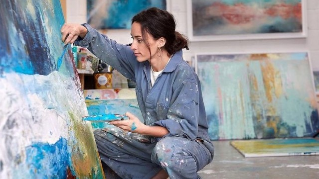 Cours de peinture. Image reproduite avec l'aimable autorisation de Red Letter Days. Femme peignant sur une toile avec de la peinture bleue, blanche et jaune, avec des tableaux en arrière-plan.
