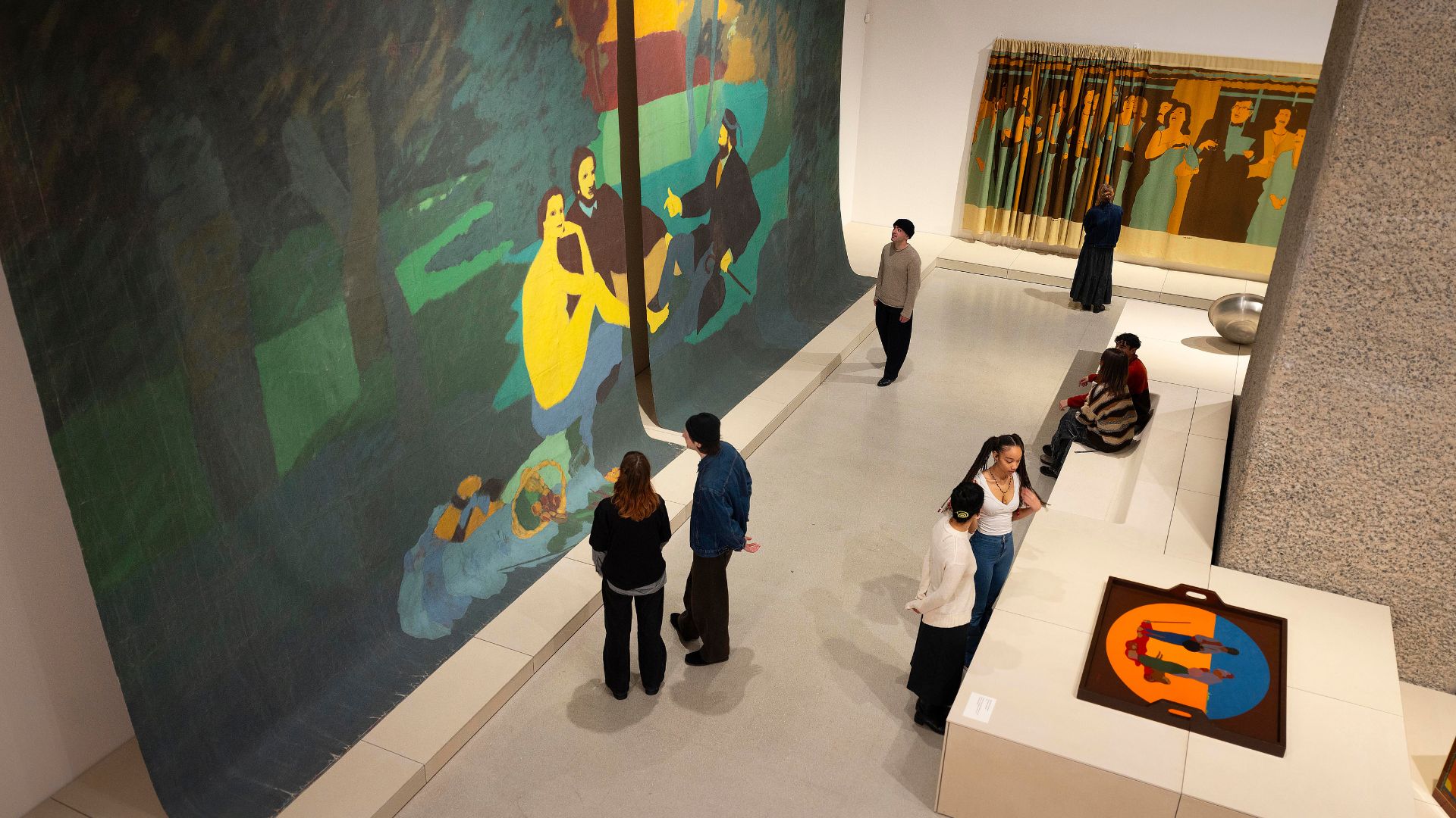 Multiple people wandering around exhibition space with colourful artworks at Barbican Art Gallery.