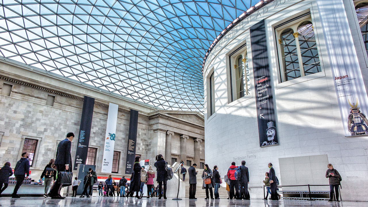 Walk under the glass ceiling of the The Great Court at the British Museum. Image courtesy of Nicolas Lysandrou/Unsplash. Visitors are walking within the Great Court at the British Museum and its blue dome.
