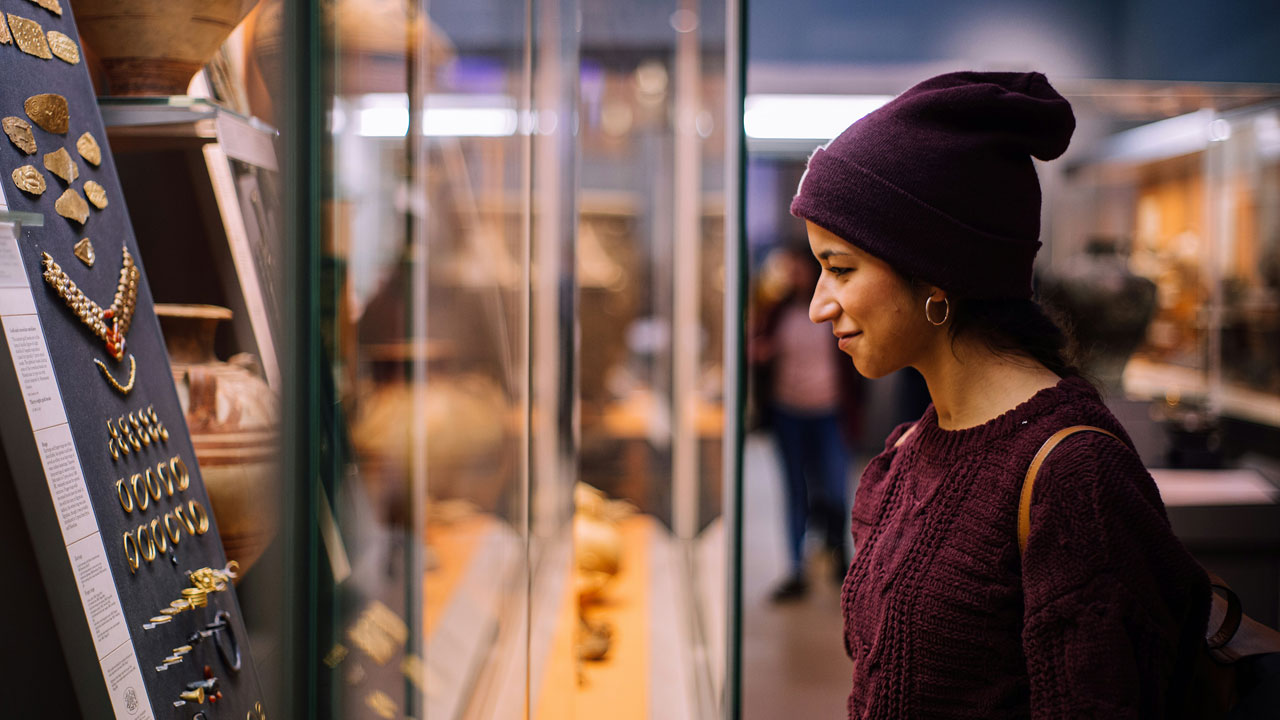 Explorez de nombreuses collections d'art au British Museum, Photo par Matteo Vistocco sur Unsplash Une jeune femme se tient devant une vitrine au British Museum exposant d'anciens bijoux.