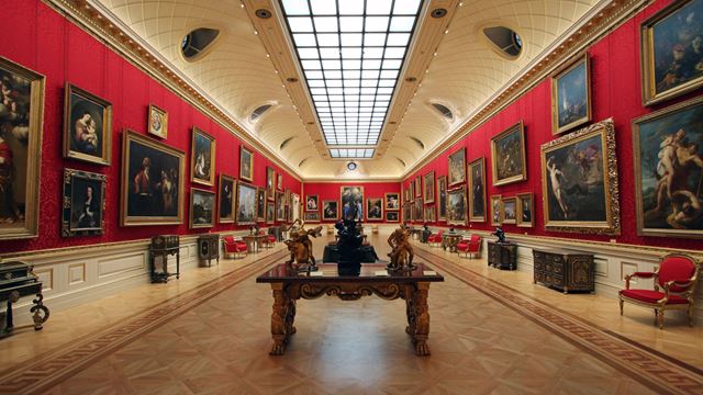 A view along the Great Gallery at The Wallace Collection, with paintings lining the walls on each side, parquet flooring and a ceiling window flooding the room with light.