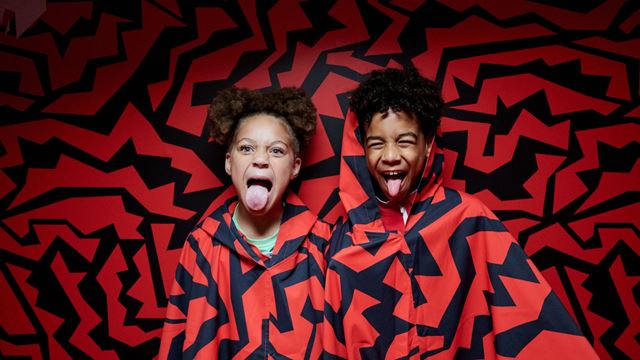 Two children pose in a red and black illusion room at Paradox Museum London, wearing matching patterned ponchos that blend into the immersive installation.