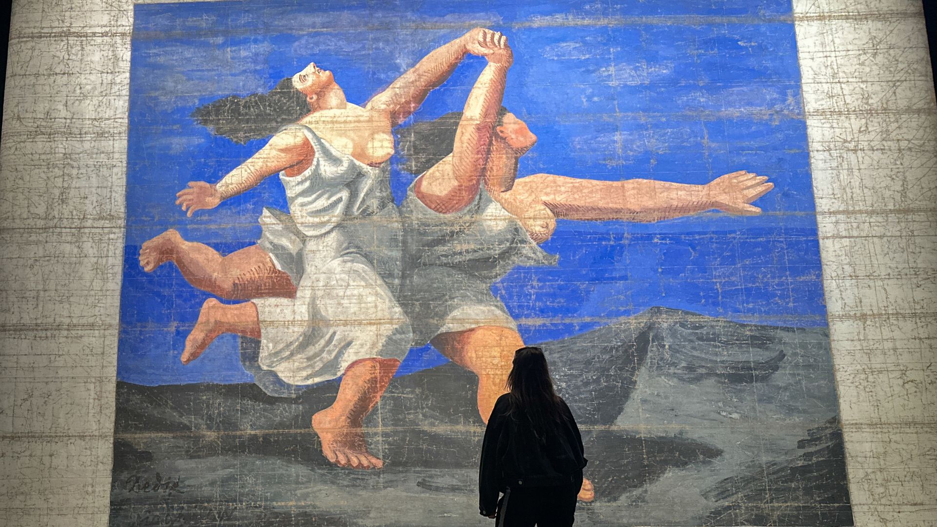 A woman stands in front of a large theatre front cloth with two dancing ladies with a blue sky.