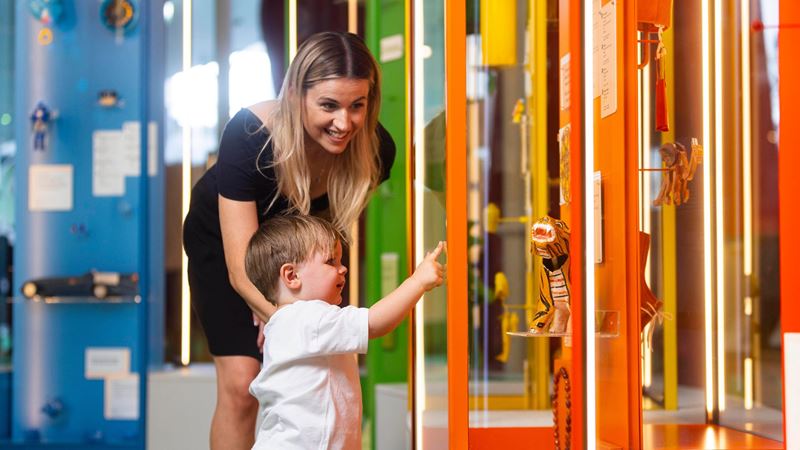 Adult and young child looking at colourful object displays in a gallery at Young V&A, Bethnal Green.