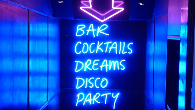In a stairwell an arrow points down to writing in neon lights reading bar, cocktails, dreams, disco, party. 
