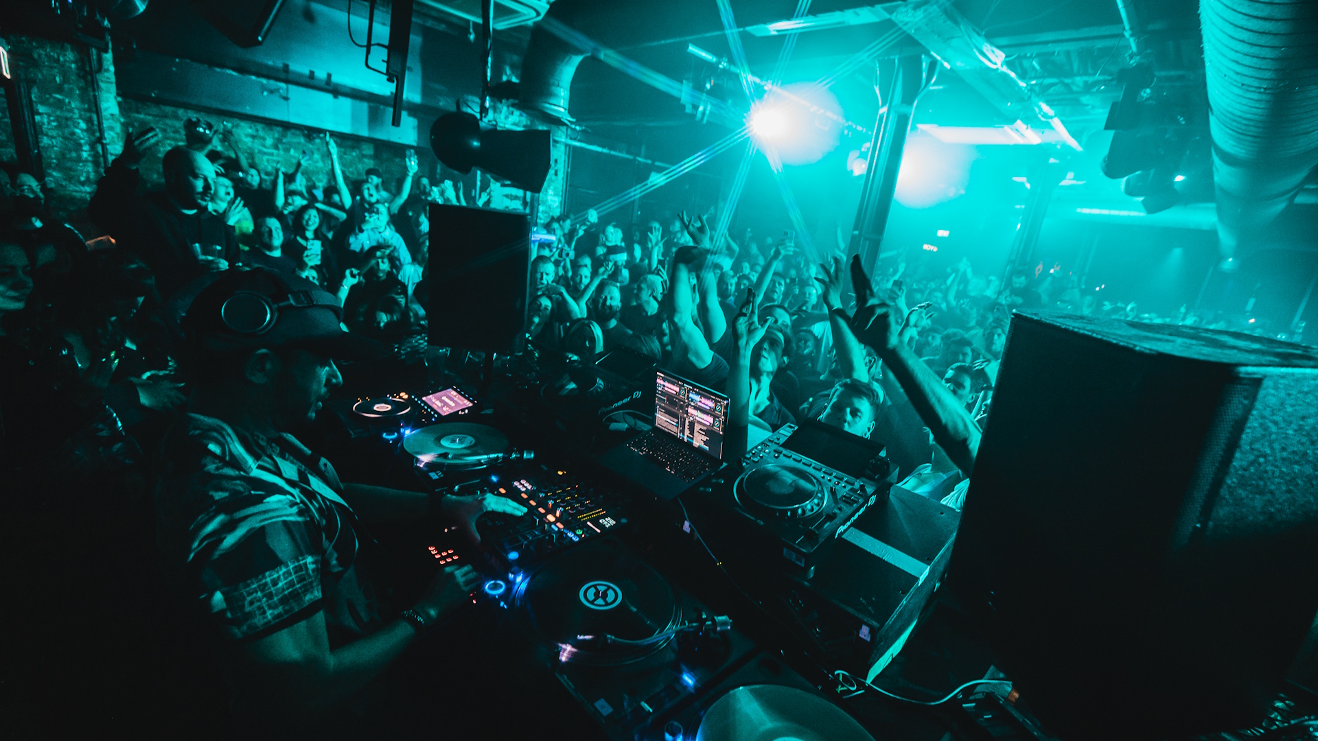 DJ spinning tunes in front of a crowd dancing with their arms up in the air in a nightclub lit up in green.