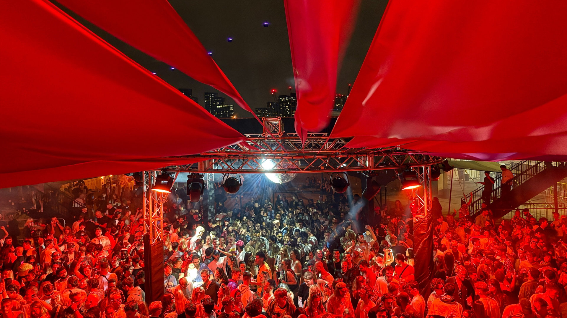 A photo of an outdoor stage at The Cause in London with a giant disco ball and red tapestry hanging over a large crowd of people