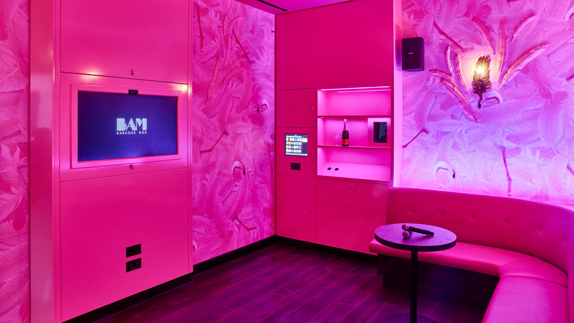 A photo of a pink karaoke booth with two microphones on a table in front of the curved sofa, a TV on the wall and a bottle of prosecco on a shelf