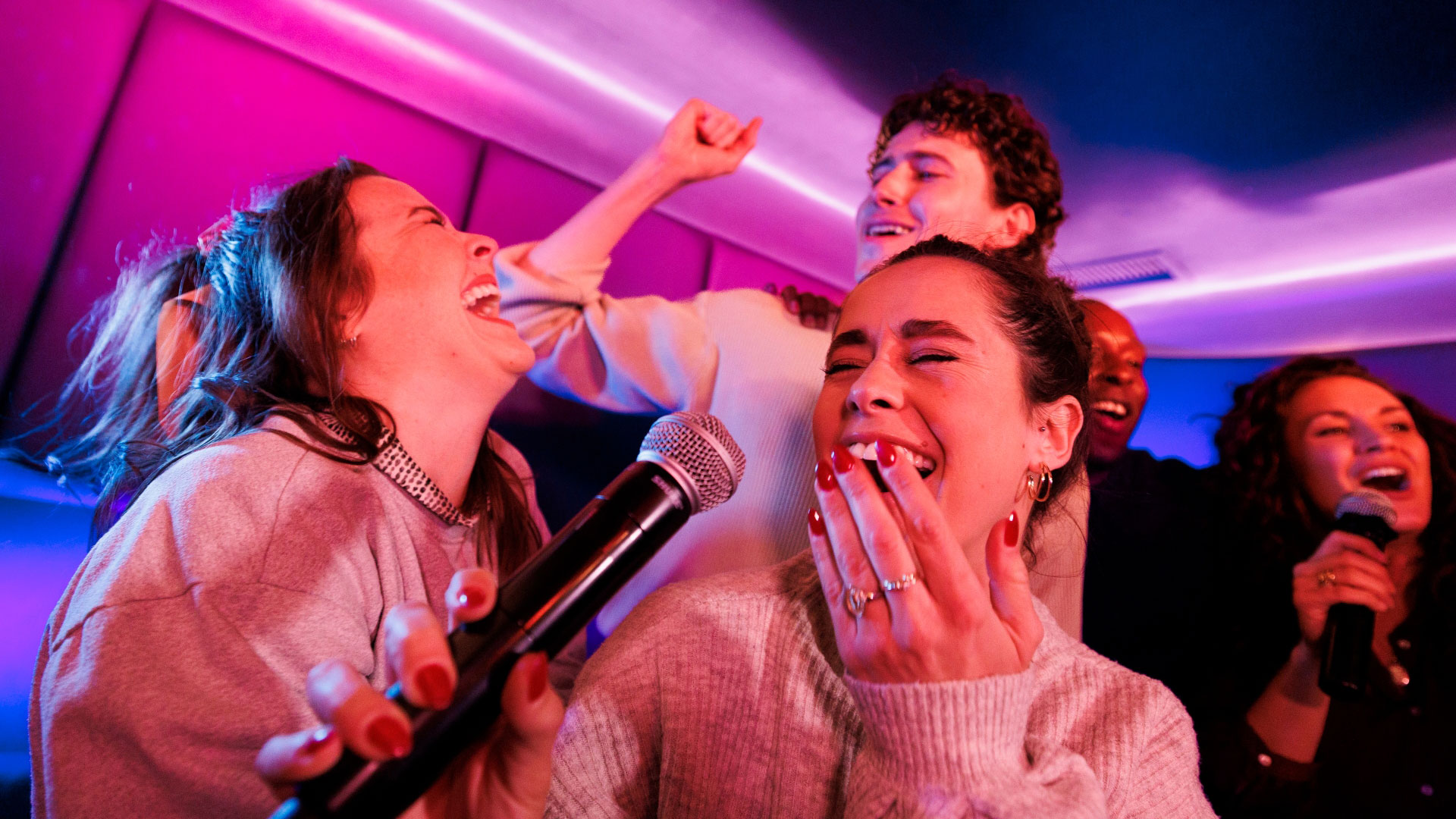 A photo of five friends laughing and singing into a microphone at Lucky Voice karaoke with pink neon lights illuminating the room