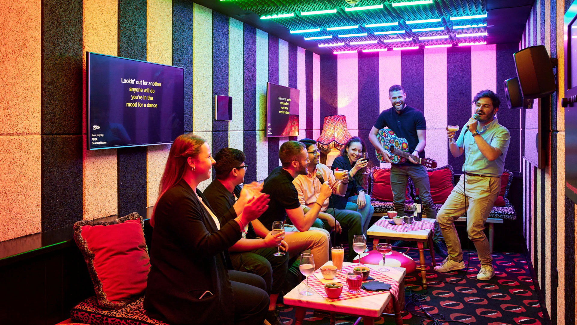 A photo of friends in a Japanese-style karaoke room at Mama Shelter, with one man stood up singing into a microphone, another man pretending to play an acoustic car and Abba lyrics on the screen whilst five friends sit on the sofas clapping and smiling