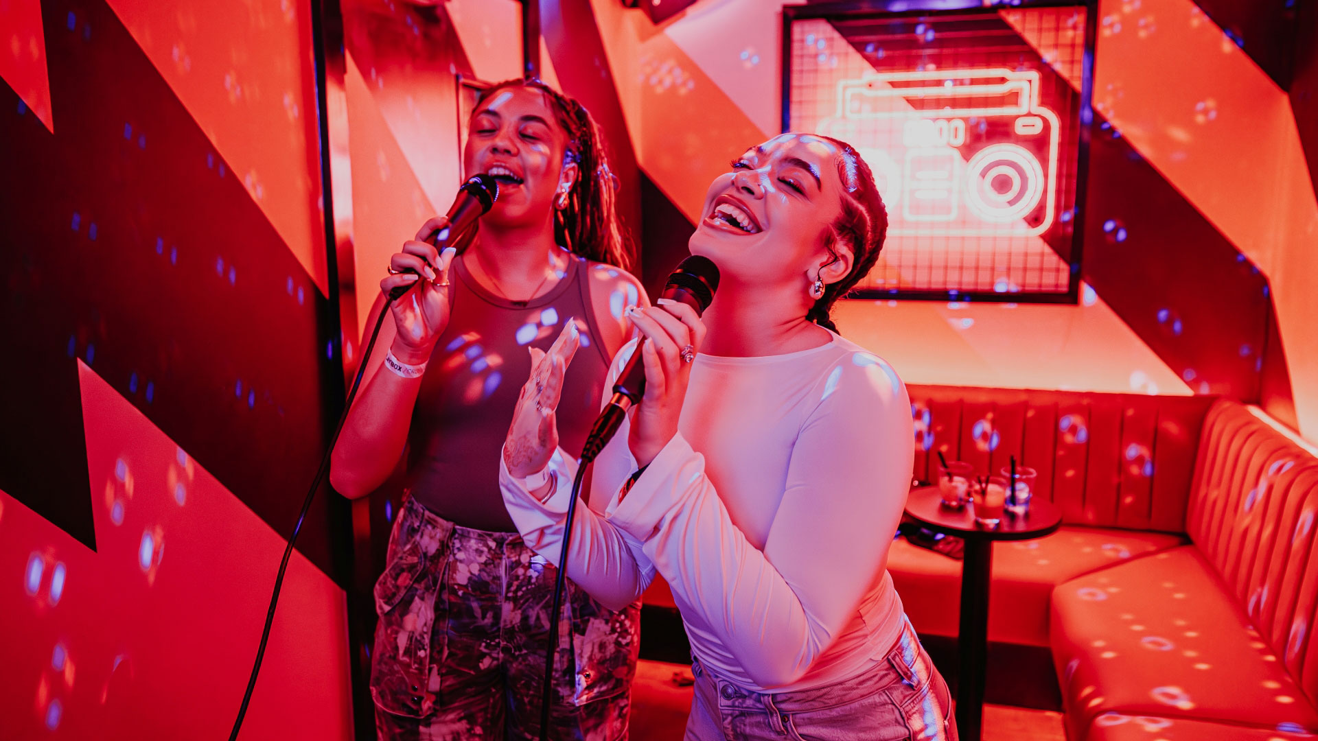 A photo of two girls smiling and singing into microphones inside a karaoke booth at Playbox with red lighting 