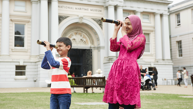 A young boy in a striped top and older girl both in a pink dress look through telescopes on the green outside of the National Maritime Museum