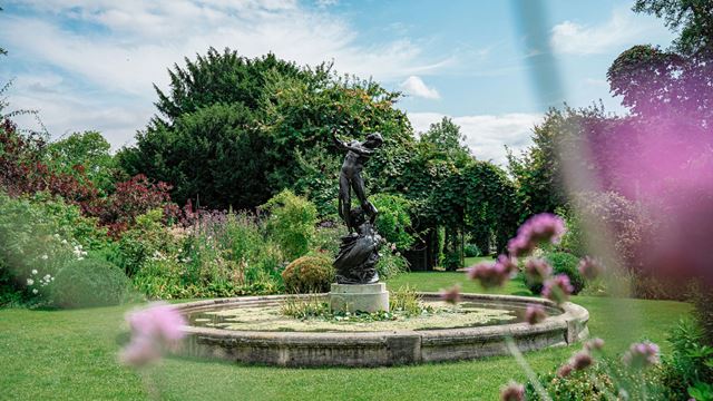 A circular stone pond with a bronze figure in the centre, surrounded by purple flowers.