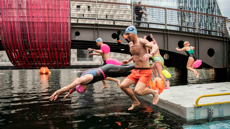Swim among the high-rise buildings of Canary Wharf in the summer months. Credit: Sean Pollock. Image courtesy of Canary Wharf Group. People jumping and diving into the water during Canary Wharf Open Water Swimming