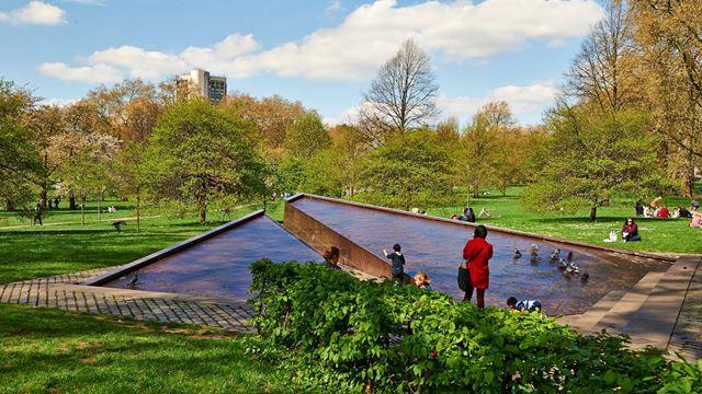 A woman and children play in the Canada Memorial, a triangular water feature made from metal, with the expanses of green space in The Green Park in the background.