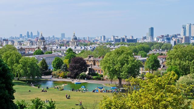 People sit on green lawns in Greenwich Park in front of a view of the London skyline.
