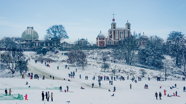 Greenwich Park in the snow © Visit Greenwich. The hill at Greenwich Park covered in snow, with many people standing around, and the Royal Observatory in the background.