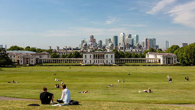 Gruppen von Menschen sitzen auf dem Hügel des Greenwich Park mit Blick auf das Old Royal Naval College und die Londoner Skyline.