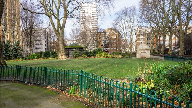 Small park with green railings and shrubs, with apartment blocks in the background.