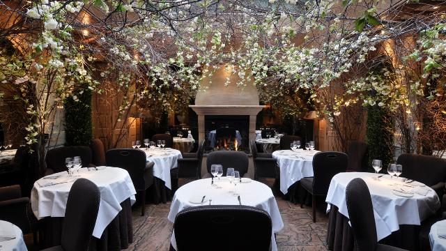 A range of beautiful dining tables with white table cloth are set by a lit fireplace in a lovely restaurant lounge with fairy lights.