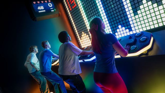 Five people next to each other in a line each with a control panel in front of them which control the light screens on the wall in front of them.