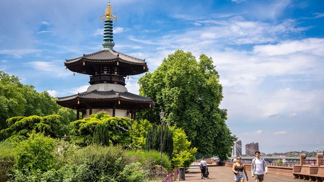 A pagoda surrounded by green shrubs and trees with a wide walkway to the right where a couple and woman with a pushchair are walking.