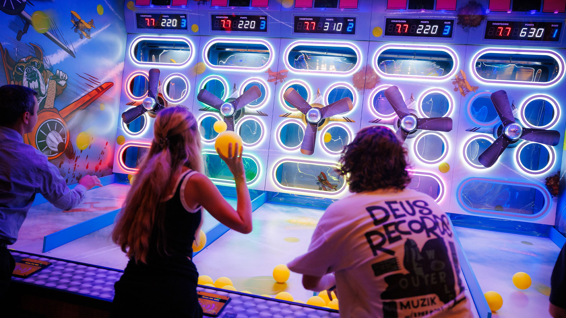 Three people try to throw yellow balls into holes which have propellers in front of them and electronic scores for each person displayed above.