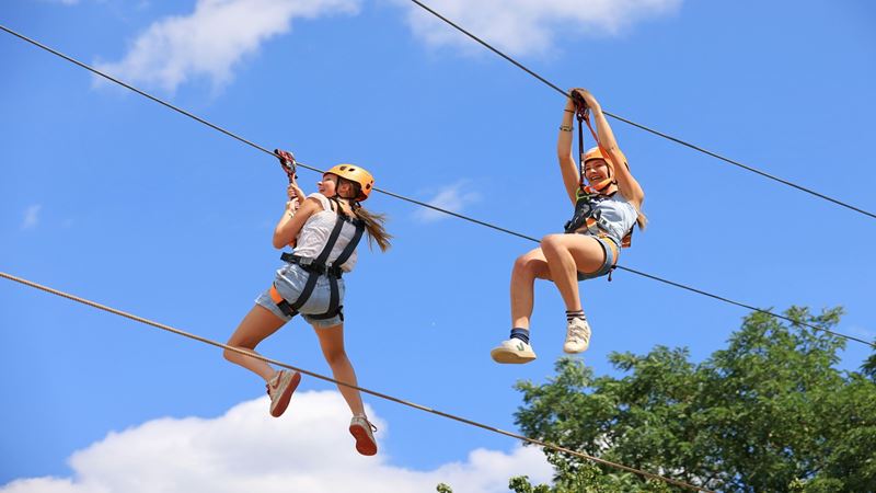 Experience thrilling activities at Gripped London. Image courtesy of Golden Tours. two girls smiling and laughing on a zip line experience, with a cloudy blue background