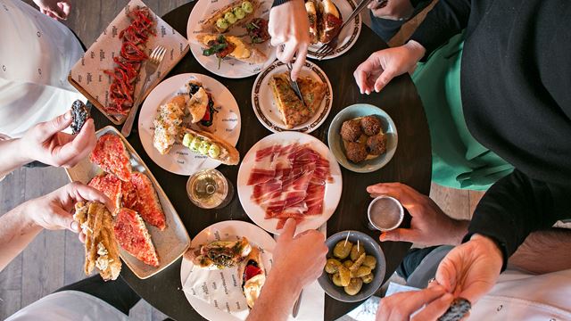 A selection of ten pica pica small plates of charcuterie, bread, olives and more shown from above on a small round table with hands reaching in for food.