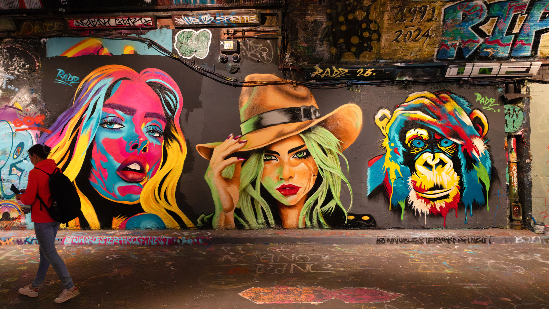 A woman walking past a wall decorated with brightly-coloured street art featuring the faces of two women and a chimpanzee.