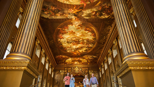 Deux couples se promènent dans le Painted Hall. Deux piliers dorés se trouvent de part et d'autre des couples au premier plan et le plafond peint les surplombe.