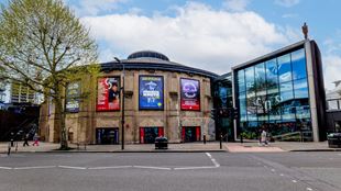 A daytime view of the Roundhouse taken from across the road with four large posters on the walls advertising upcoming events.