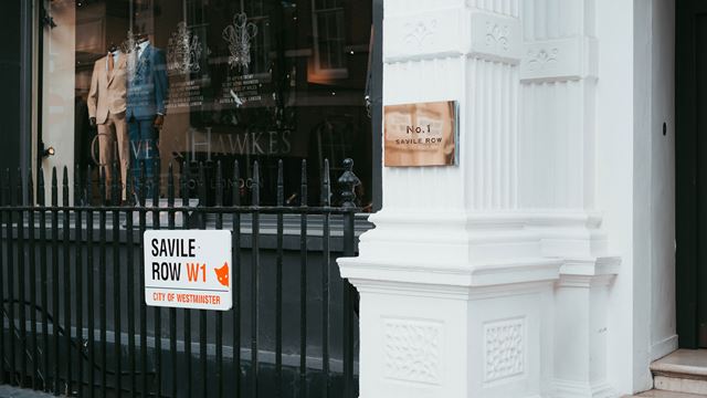 A white pillar and black railings at the entrance to a shop with suits in the window, the pillar and railings display signs saying Savile Row.