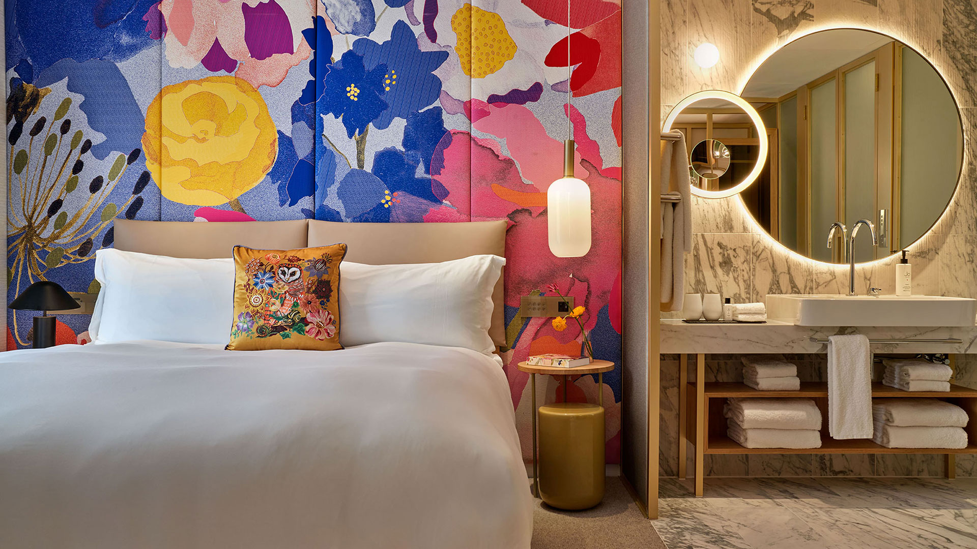 Stay in a spacious Lane Suite at The BoTree London, Curio Collection by Hilton. Image courtesy of Hilton. A large white bed with a colourful floral scene on the back wall behind it, next to a marble bathroom and large circular mirror