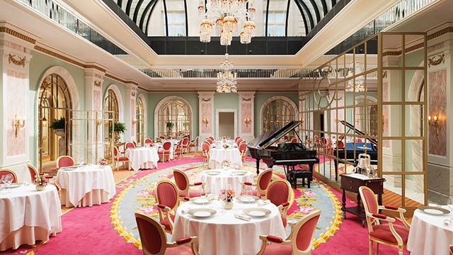 A round table set for dining in the pink and pale green restaurant with a glass ceiling, chandelier and grand piano.