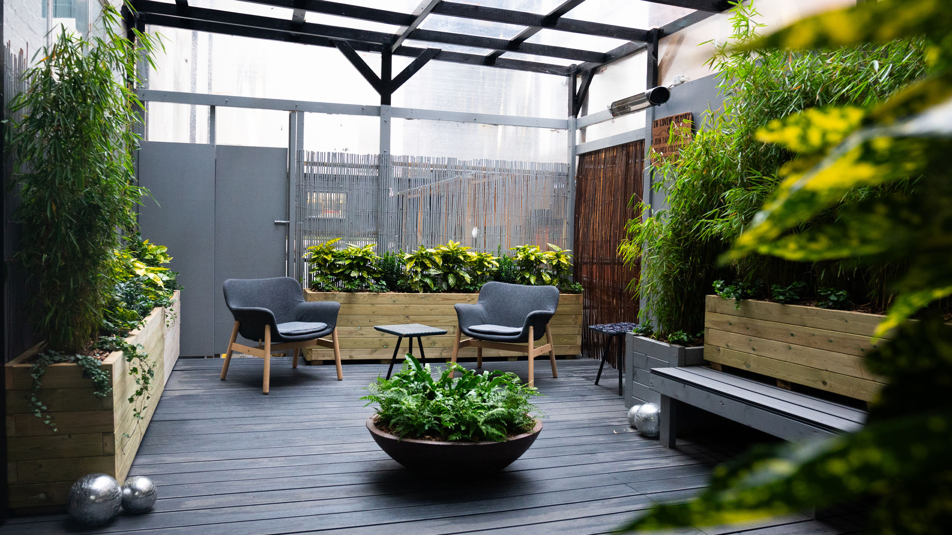 Grey seats and benches in a covered courtyard lined with green plants in wooden troughs and a bowl of ferns in the middle of the space.