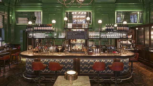 Red bar stools in front of the bar which sits in the middle of a room with wooden parquet flooring and green painted walls.