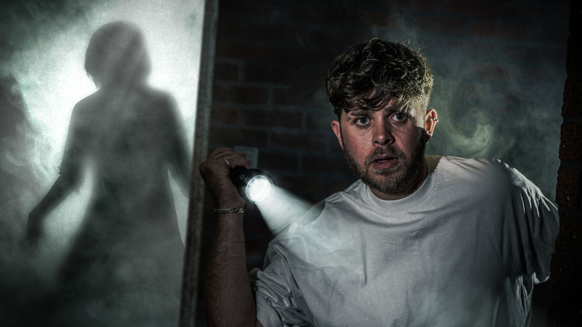 Man in grey hoodie holding a torch and looking at a reflection of a ghostly figure.