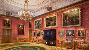A rust red drawing room adorned with gold framed paintings and a large chandelier at windsor castle.